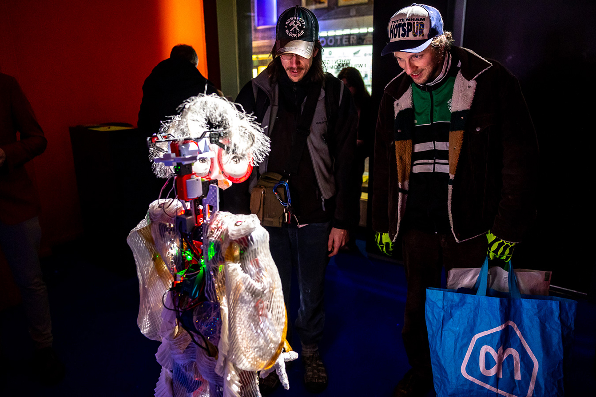 The audience is watching 125 cm tall robot standing on a platform in a dark gallery space. The robot is talking to them. In the chest of the robot is green light.