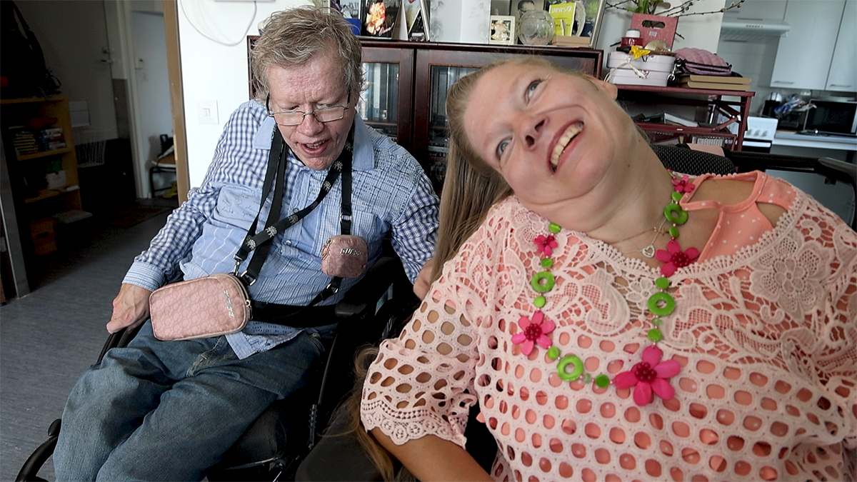 A couple is sitting in their wheelchairs in a living room. A man is combing his wife's long blonde hair.