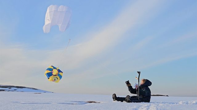 Jenni-Juulia sits on the snowy tundra, flying a yellow kite marked with the ISA symbol.