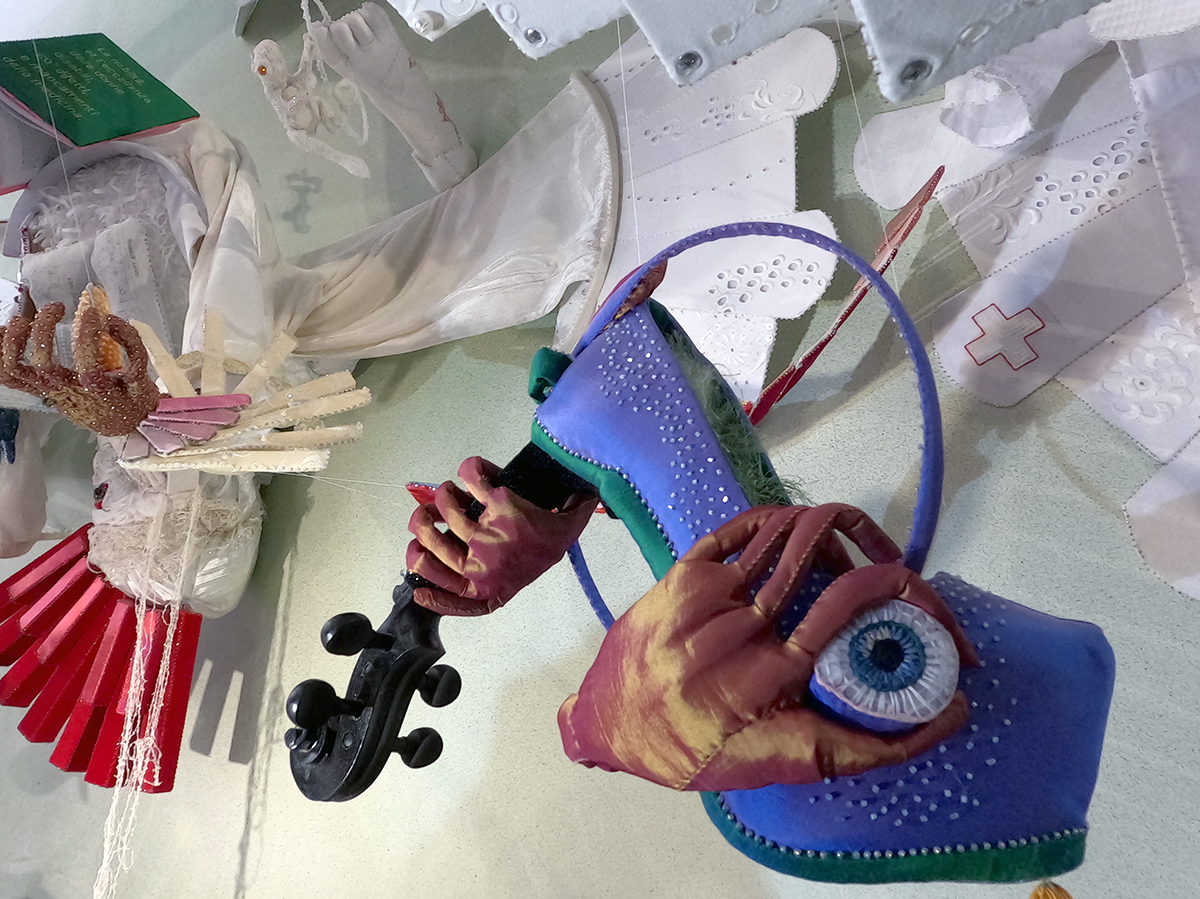 Blue taffeta high heel with beads and an eye has the neck of a violin as the heel. A textile hand has turned fingers around the eye.