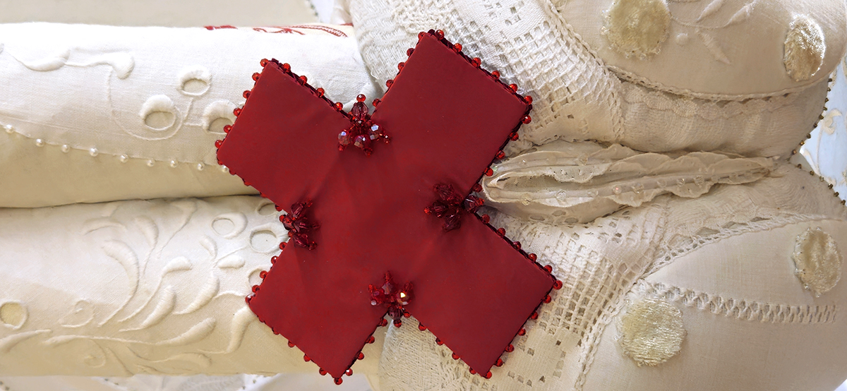 White fabric bum with a red cross in front of it. Beading, red chrystal beads and white-on-white embroidery,
