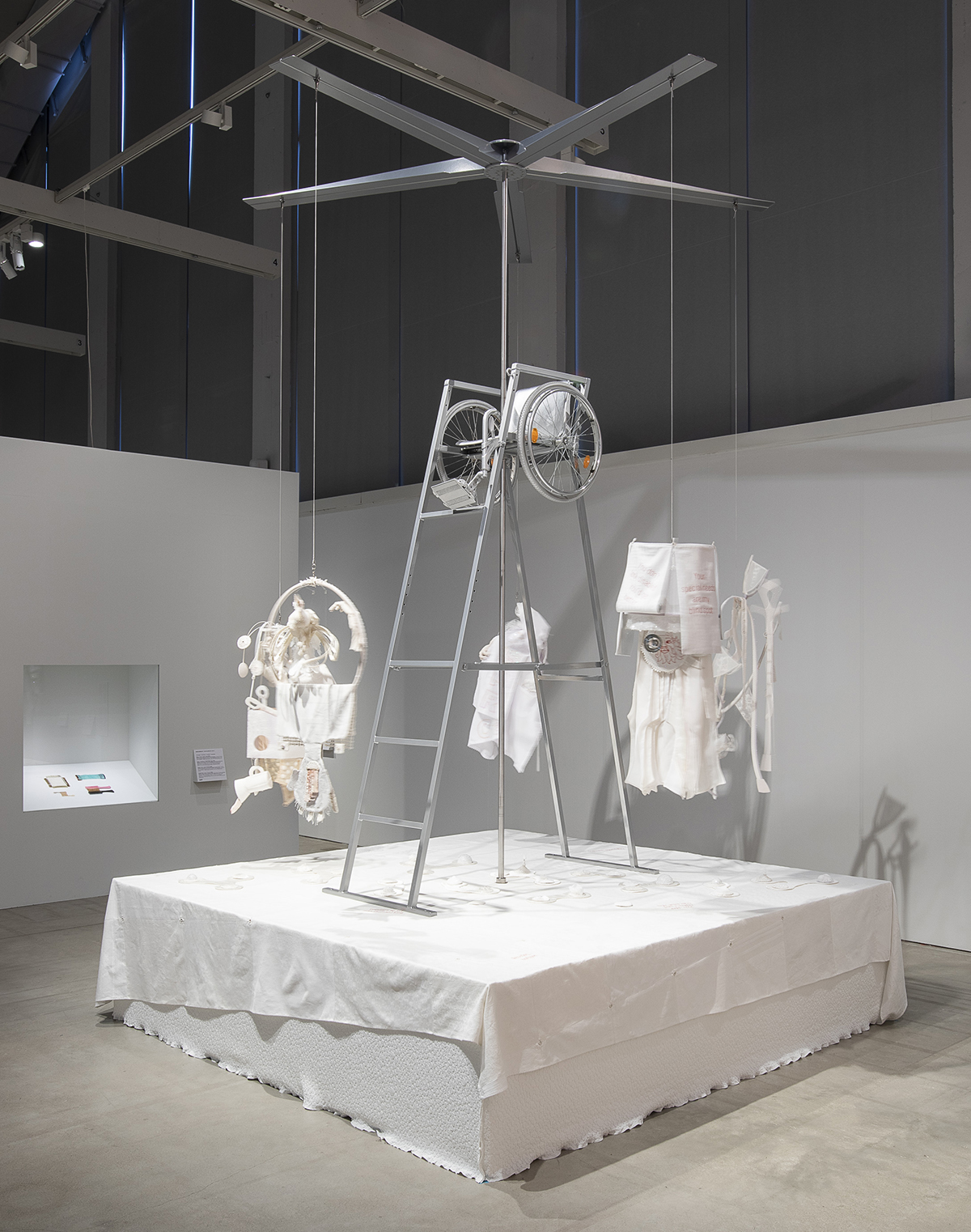 On a 3 x 3 meters pedestal covered with an enormous cloth stands a tennis judges chair. Its seat is replaced by a silver colored wheelchair. A pole rises trought the wheelchair and it is topped with a massive helicopter propeller. Hanging from the propeller blades are six textile sculptures made of fabric. There are human and animal figures, ableist texts, crutches and clothing.
