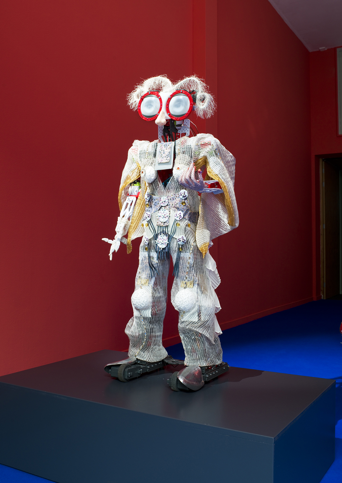 Robot standing on a pedestal in a red gallery room at 1646 Experimental Art Space.