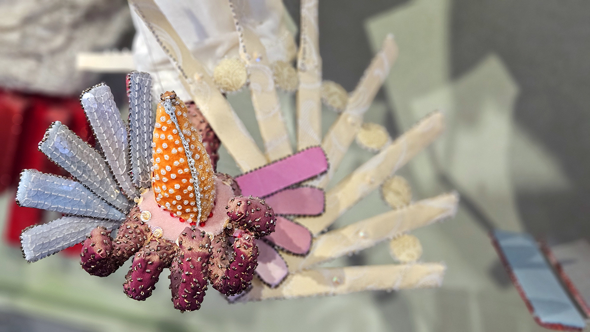 Textile hand with tiny beads. From the wrist portrudes blue, pink and light yellow wings. In the center of the hand is a large bird's beak.