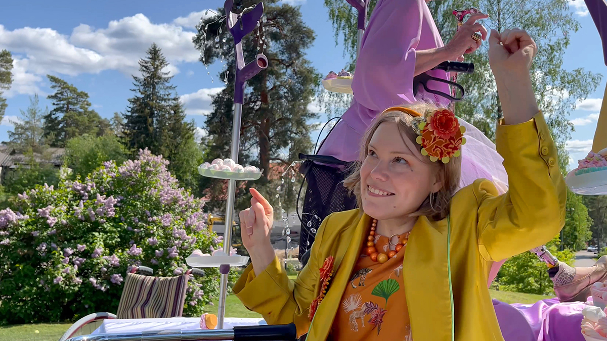 Poet Sanni Purhonen sitting on a table dancing in a yellow silk jacket. Behind her is a cookie stand made from a forearm crutch.