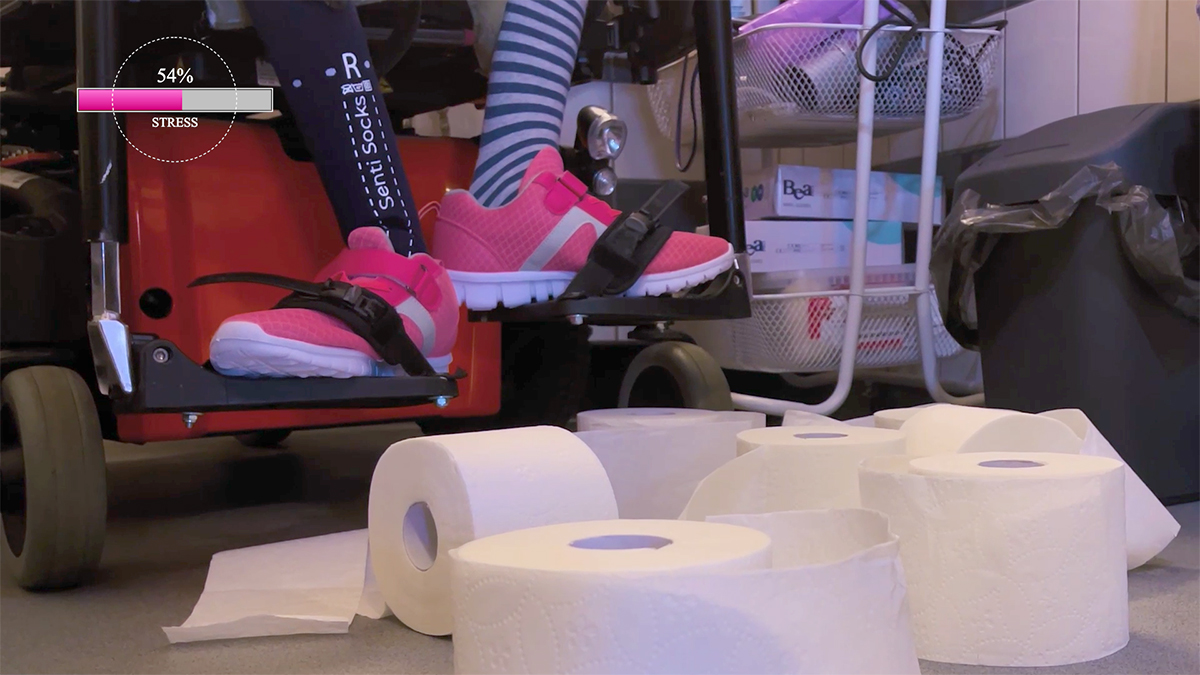 Toilet paper rolls on the bathroom floor. Behind them, there are woman's legs on a wheelchair footrest. On one foot, she wears a blue sock that measures her stress.