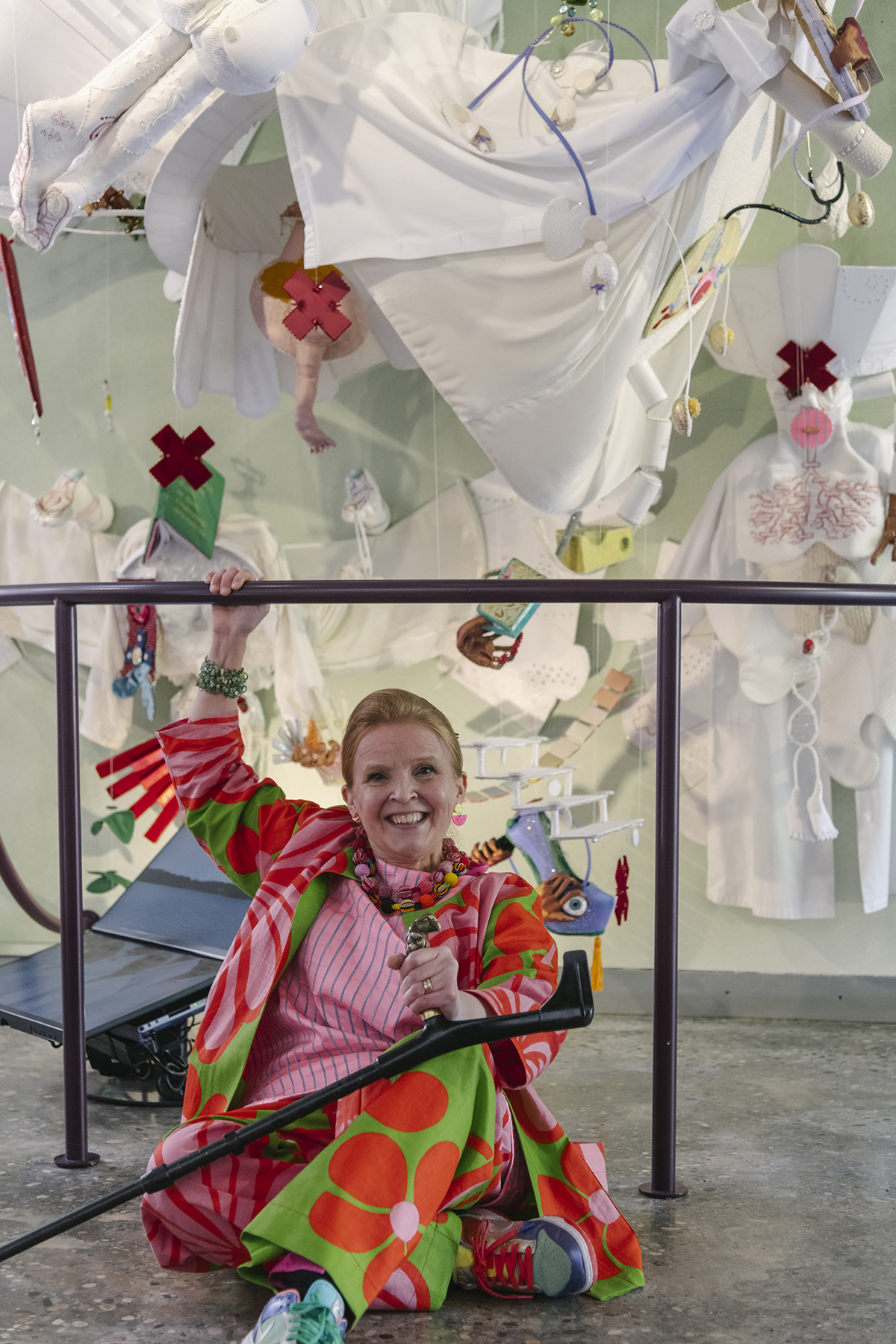 Jenni-Juulia sitting in a colorful print outfit on the floor with a crutch. Behind her is a large light colored textile installation with multiple embroidered details hanging from the ceiling.